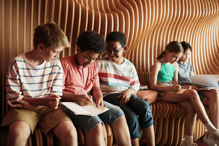 A Group Of Kids Sitting On A Wooden Seat Writing On Notebooks