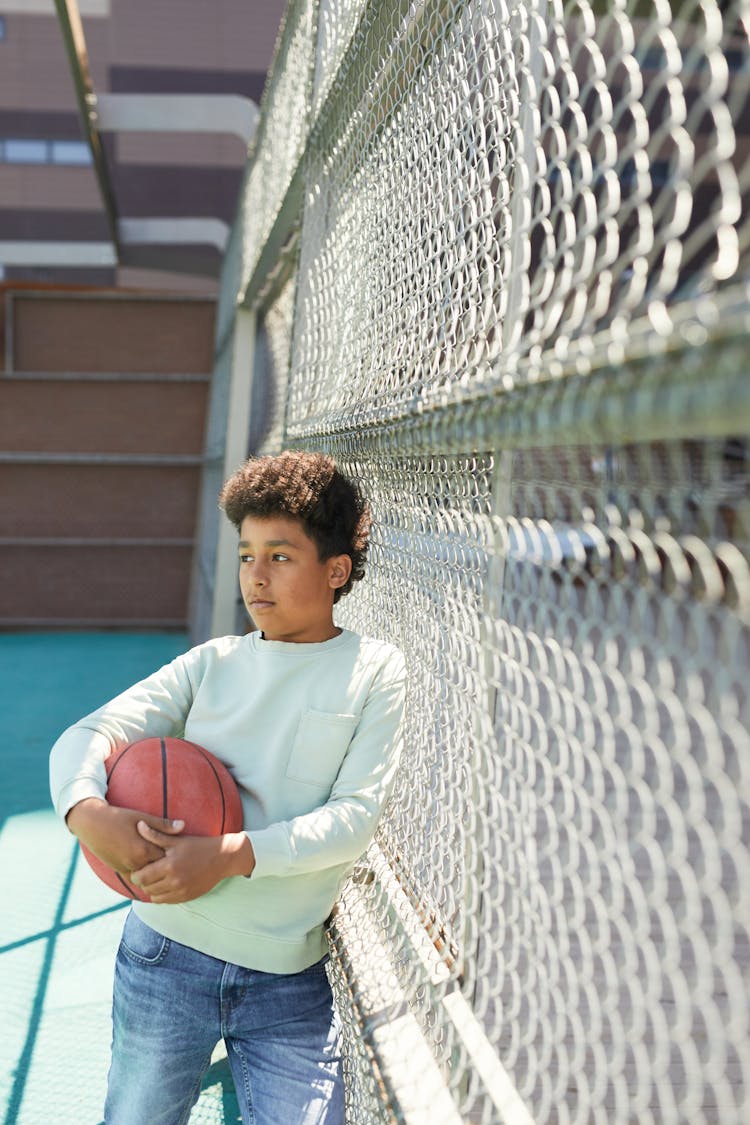 Kid Holding A Ball While Leaning On Wire Fence