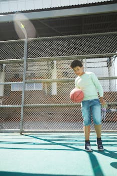 Teenage boy holding a basketball on an outdoor court, sunlight highlighting the scene.