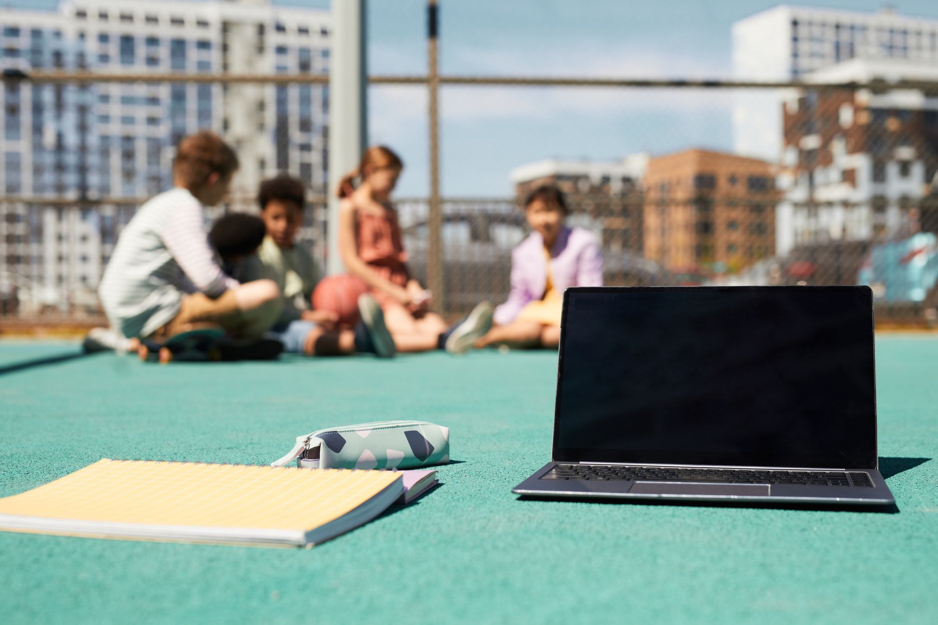 Children sitting on a sports ground with a laptop and notebook, urban backdrop.