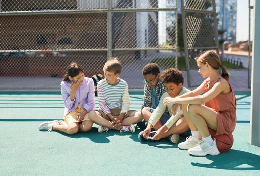Five diverse children sitting and interacting on an outdoor playground, enjoying leisure time.