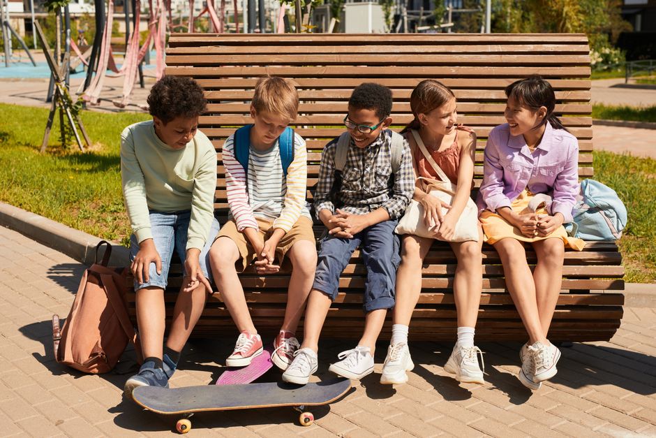 Diverse group of children sitting on a park bench with backpacks and skateboard. Sunny day showcasing friendship and leisure.
