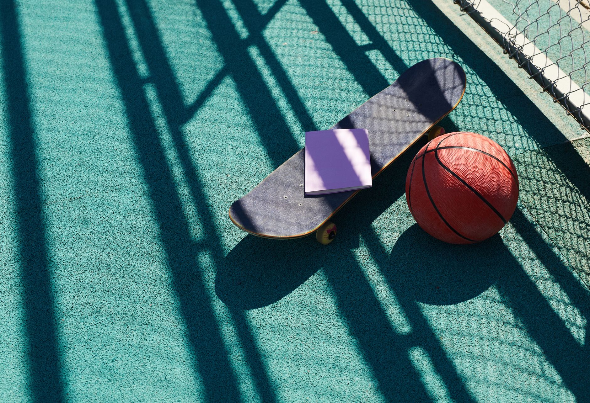 A skateboard, basketball, and notebook placed on a sunny court with shadows.