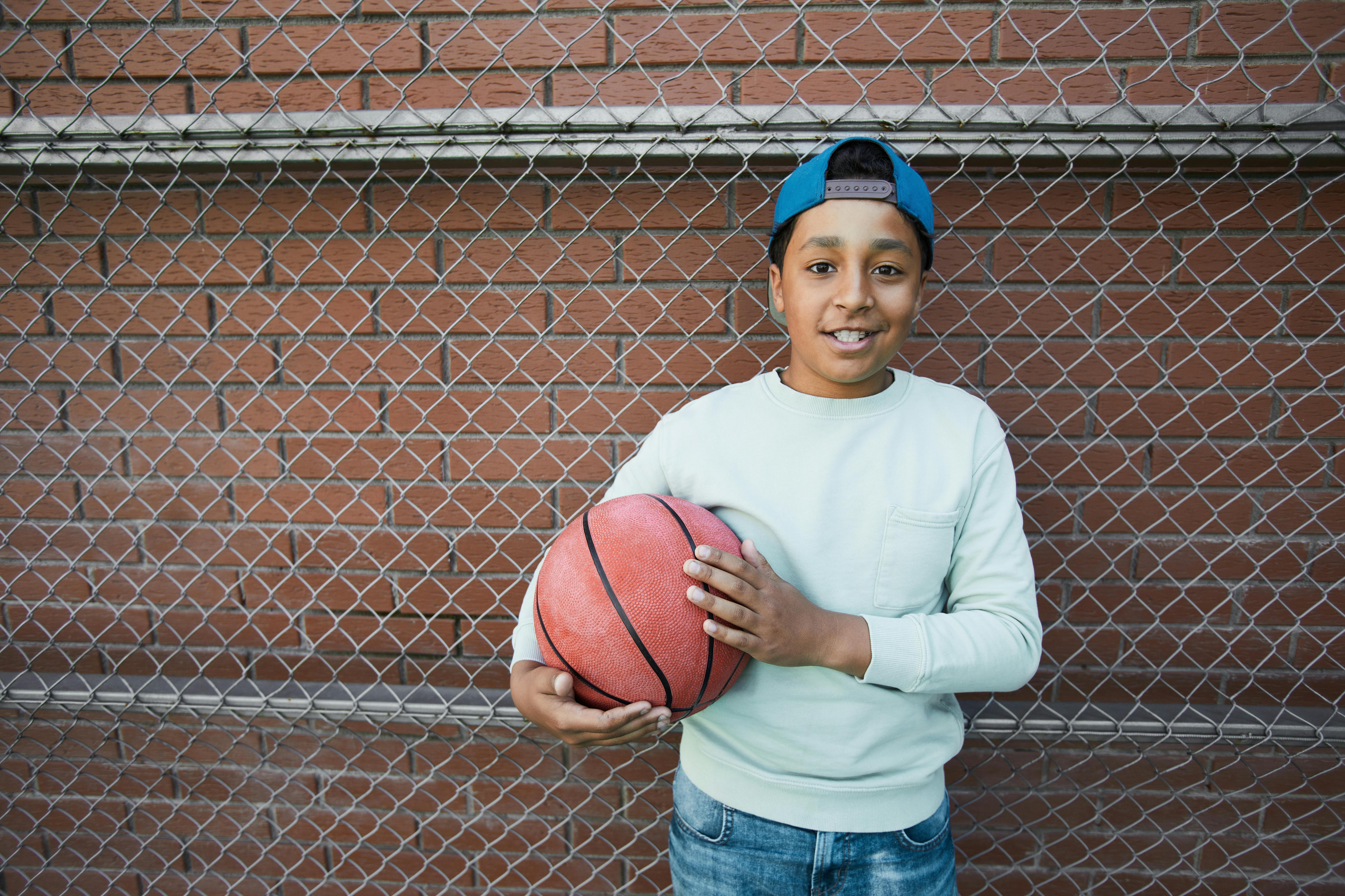 a boy standing beside a chain link fence holding a basketball