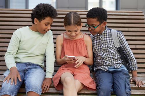 Free Three kids sitting on a bench, interacting with a smartphone, depicting friendship and technology. Stock Photo