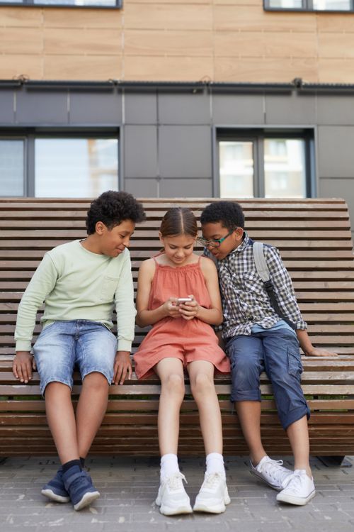 Free Three kids sitting on a bench, sharing a smartphone, representing friendship and technology. Stock Photo