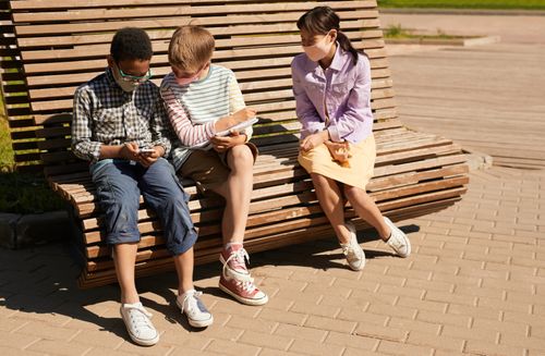 Free Three kids sitting outdoors on a bench, using tech devices, wearing face masks, enjoying a sunny day. Stock Photo