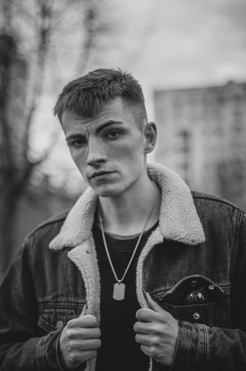Free Black and white portrait of a young man wearing a denim jacket and necklace outdoors. Stock Photo