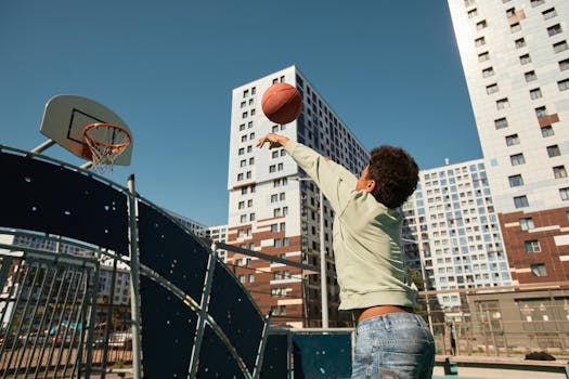 A child playing basketball in a city playground surrounded by tall buildings, enjoying a sunny day.