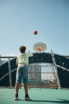 Teenage boy shoots basketball towards hoop in urban playground setting.