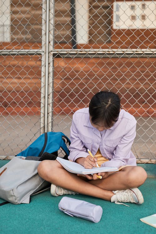 A Girl Sitting on the Ground while Writing on Her Notebook