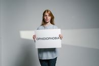 A young woman stands indoors holding a sign about ophidiophobia, conveying anxiety and isolation.