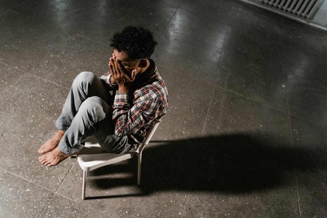 A man sitting alone in a dim room, head down, showing signs of low confidence and stress
