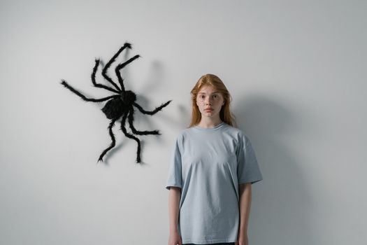 Woman stands with a large artificial spider on wall, portraying fear or anxiety.