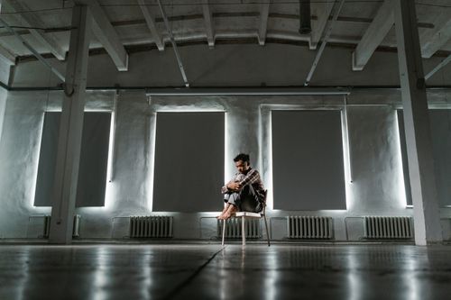 Free A lonely man sitting on a chair in a dimly lit industrial room, embodying solitude. Stock Photo