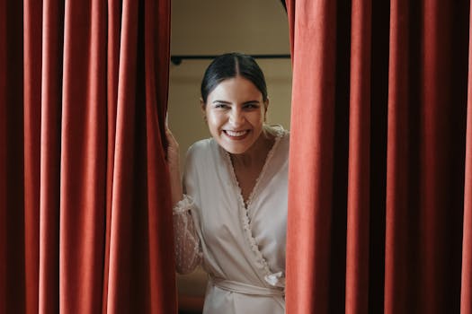 A happy bride in a white dress fitting, peeking through red curtains indoors.