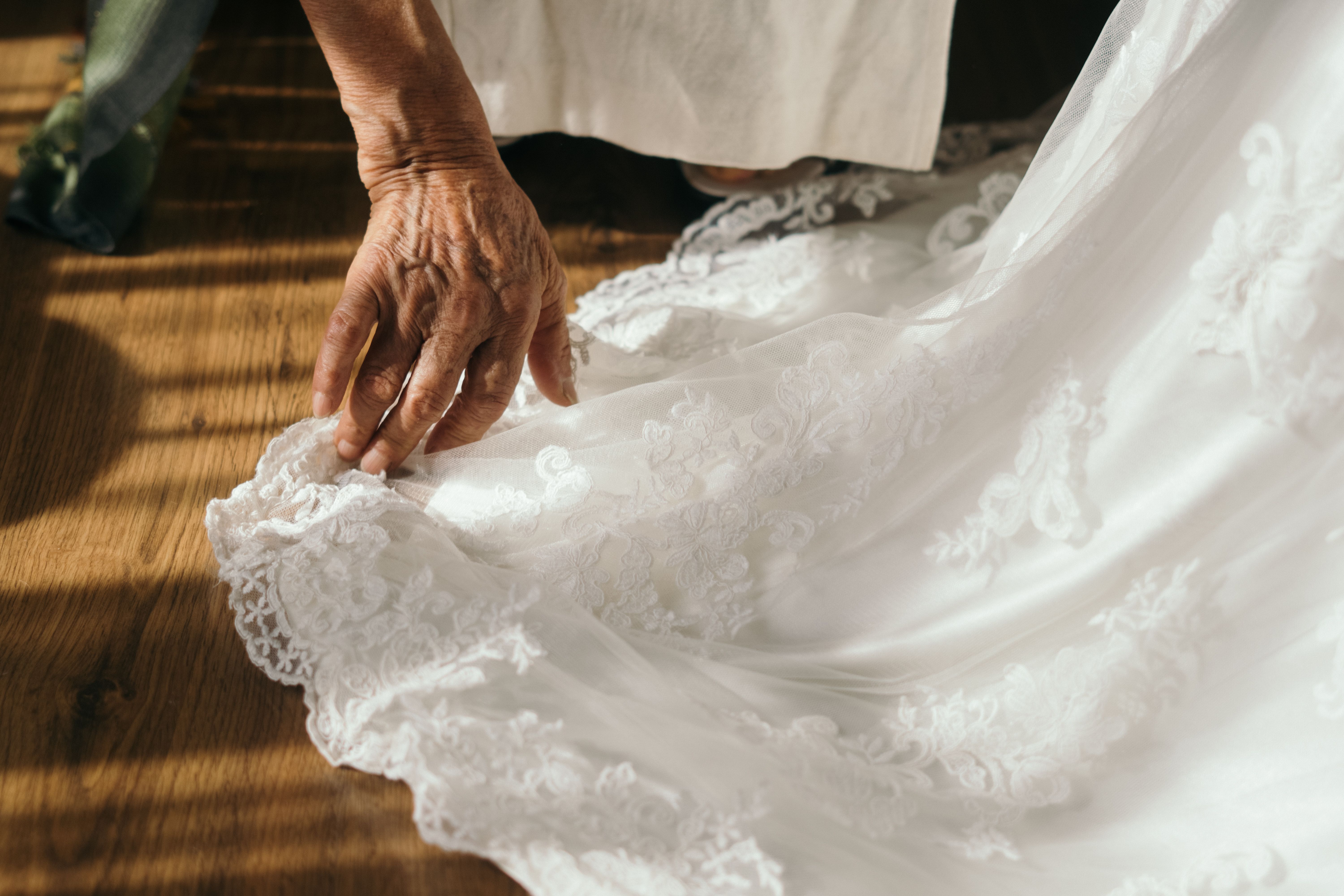 A senior adult hand gently adjusts delicate lace on a wedding dress in soft lighting.