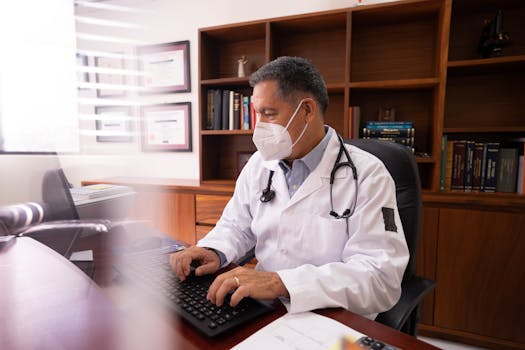 Medical professional in white coat with mask typing at desk in office setting.