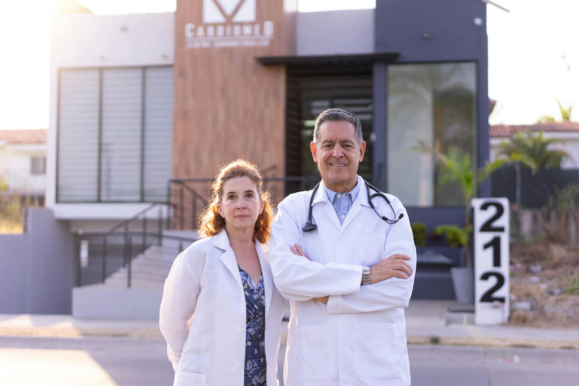 Man and woman doctors in lab coats standing confidently outside a modern medical facility.