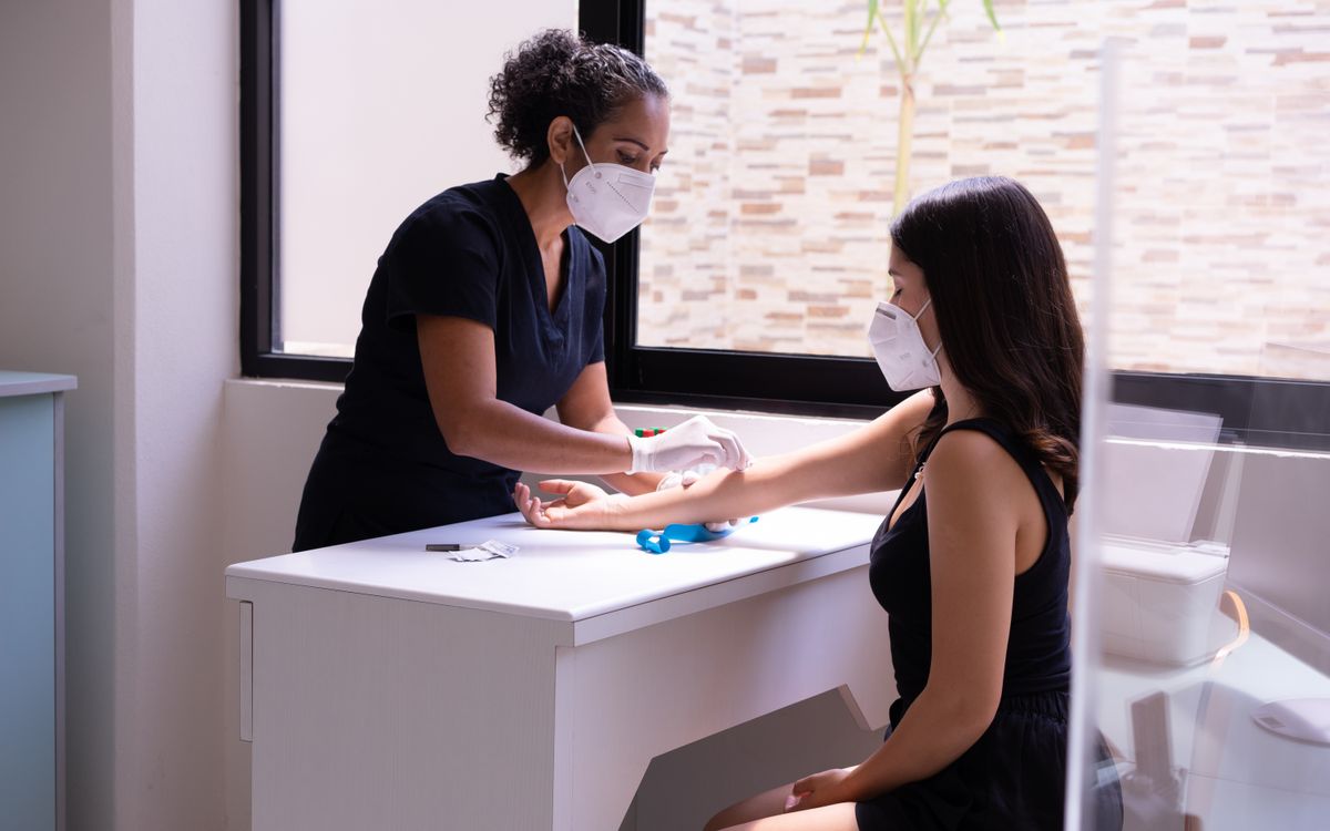A healthcare professional wearing a face mask performs a blood test on a female patient in a clinic.