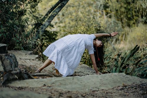 Free Woman in white dress practicing yoga on stone path in a lush forest setting. Stock Photo