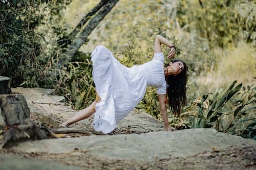 Free A woman in a white dress performs a yoga pose in a tranquil outdoor setting surrounded by nature. Stock Photo