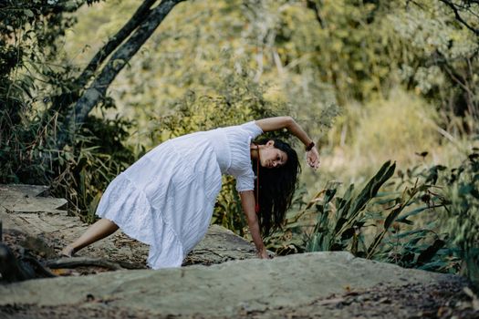 A woman in a white dress performs yoga outdoors surrounded by green forest foliage.