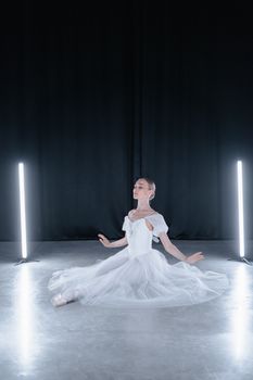 Ballerina in white dress gracefully sits on stage, lit by studio lights, embodying elegance and poise.