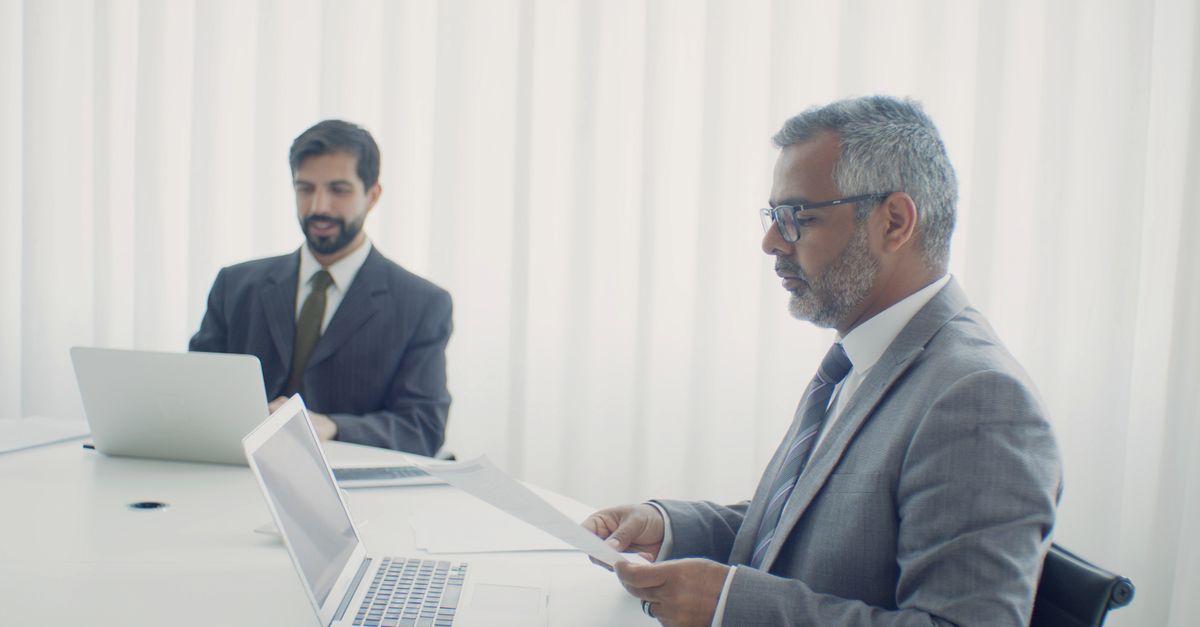 Dos colegas en un entorno corporativo, trabajando en laptops y documentos.