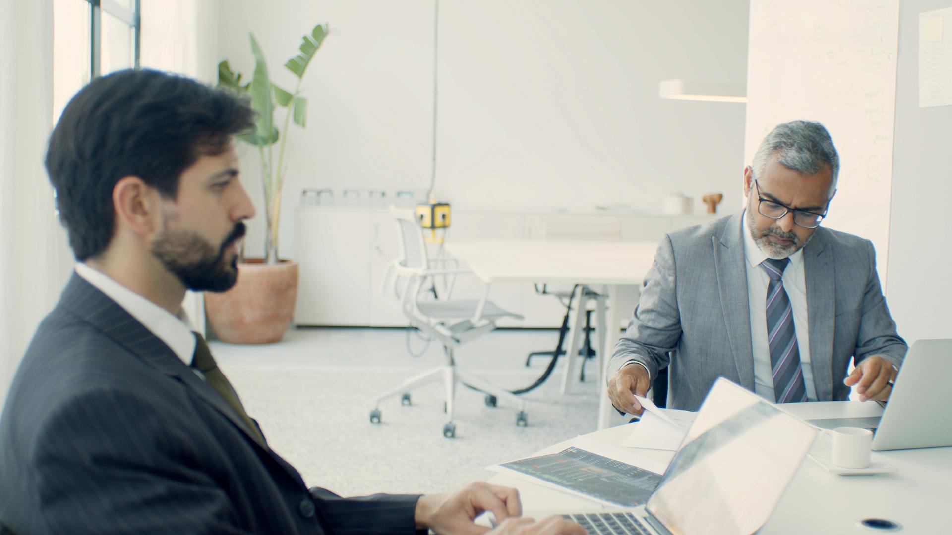 Two businessmen in formal suits working on laptops in a modern office setting.