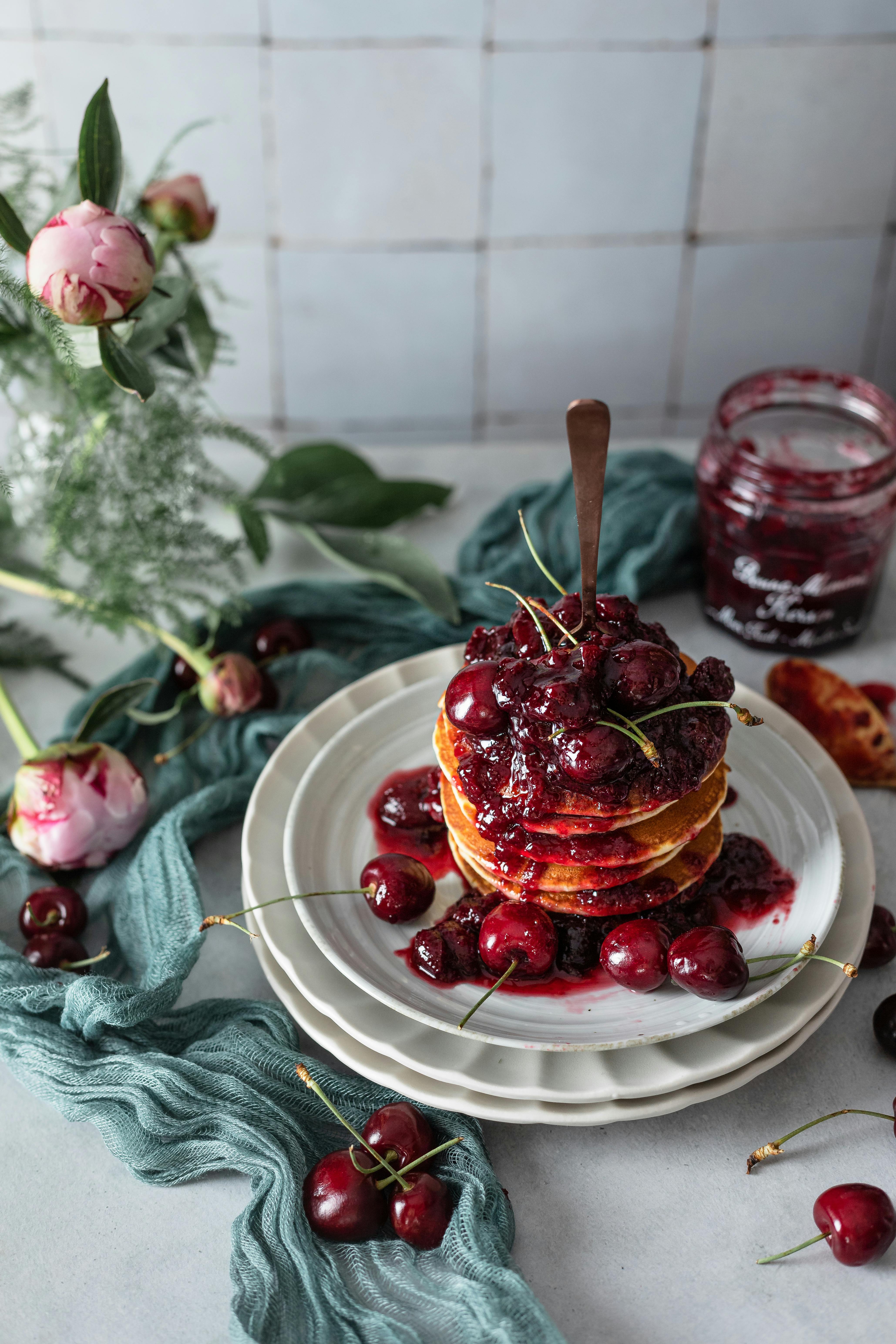 Homemade pancakes topped with syrupy cherries, surrounded by flowers on a rustic table.