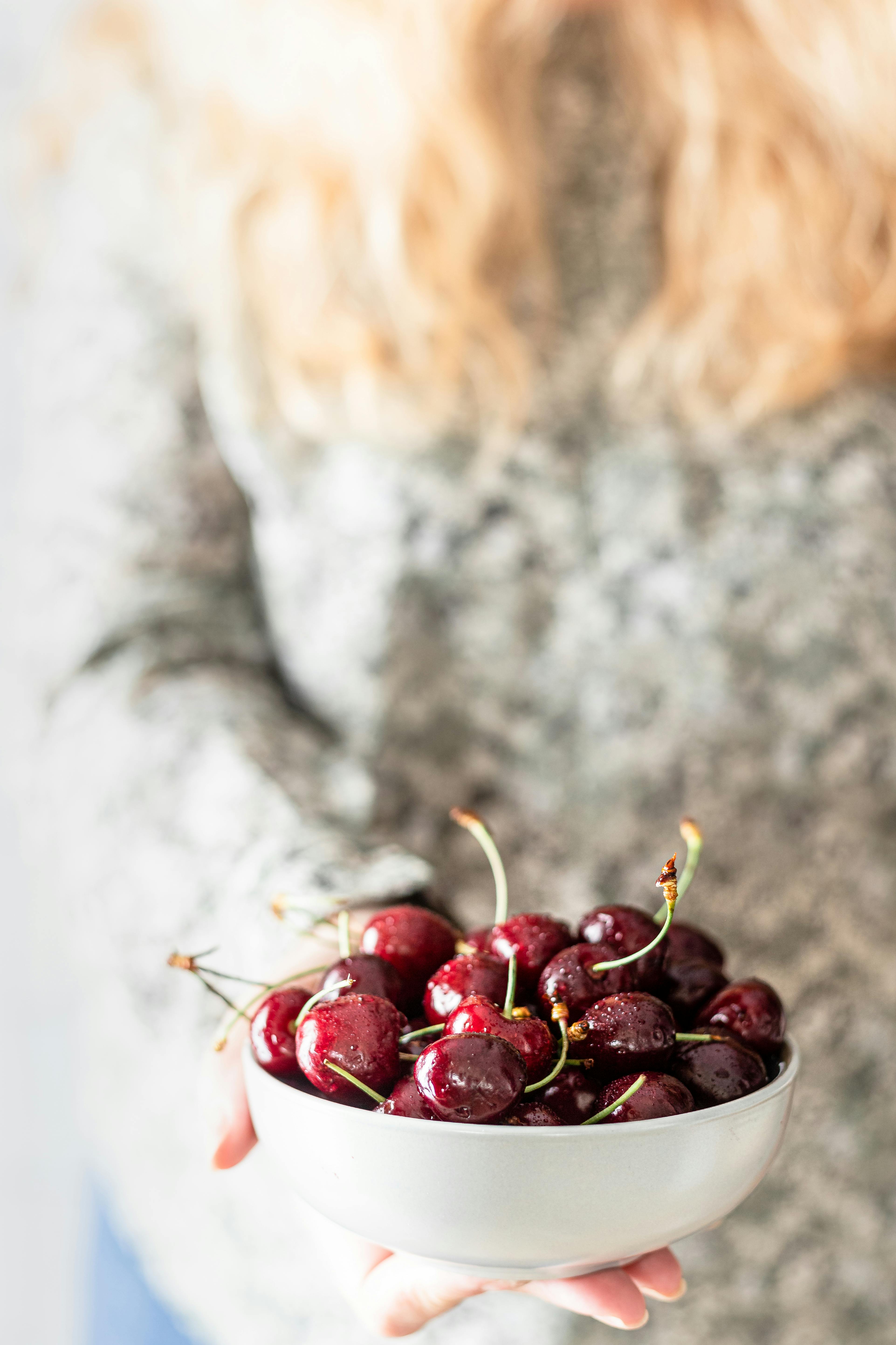 Close-up of a hand holding a bowl filled with fresh ripe cherries against a soft background.