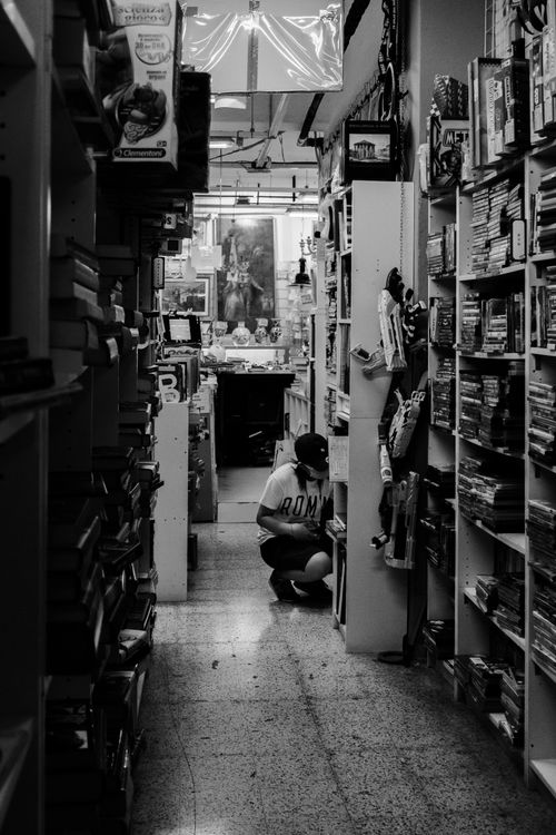 Free Black and white shot of a woman crouching in a narrow bookstore aisle lined with shelves. Stock Photo