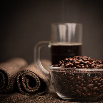 A detailed close-up image of coffee beans in a bowl with a glass mug of espresso in the background.