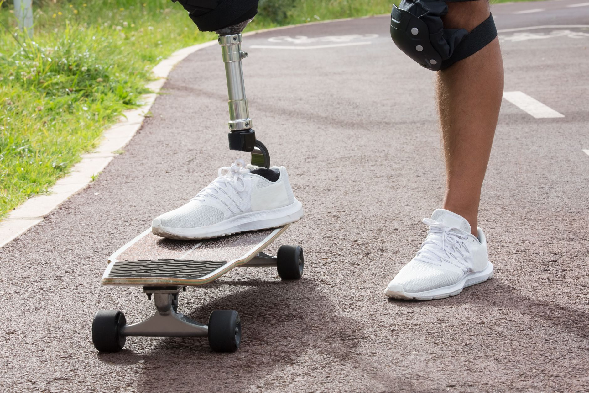 Close-up of a skateboarder with a prosthetic leg and white shoes on a paved path outdoors.