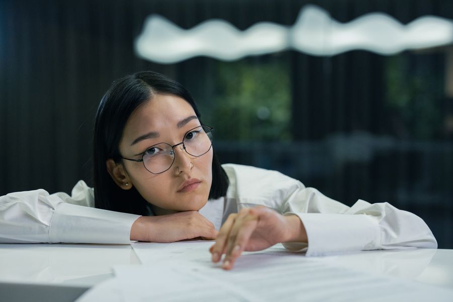 A contemplative Asian woman wearing eyeglasses and writing at a desk in an office setting.