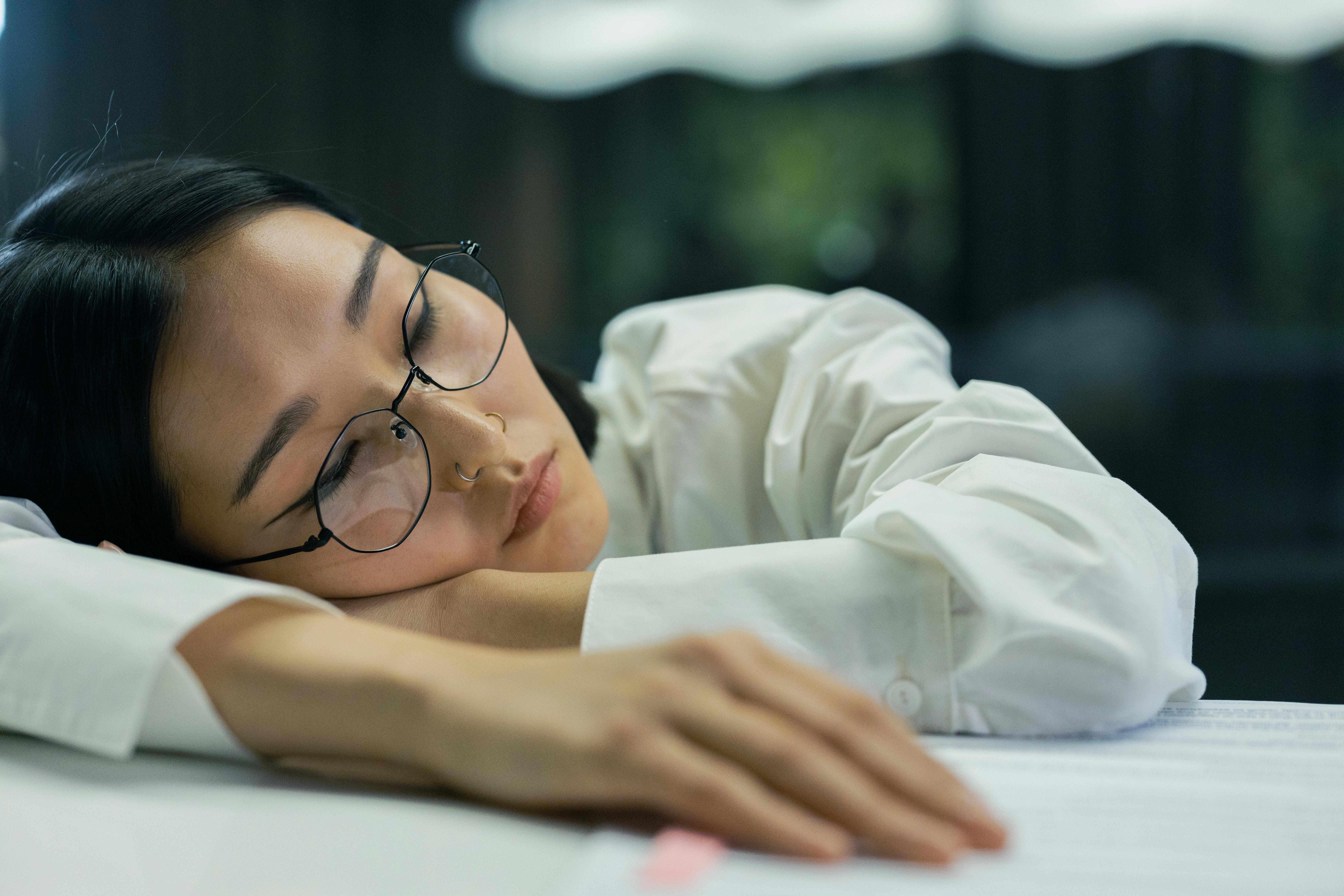 A tired woman with eyeglasses sleeps on a desk indoors, highlighting exhaustion.