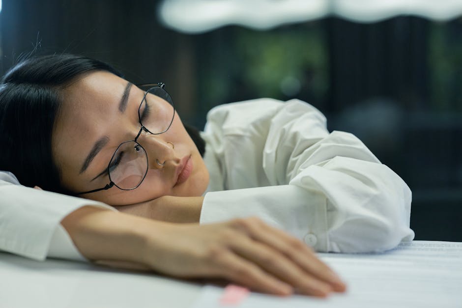 Foam vs Silicone vs Custom Earplugs: Which Best Blocks Noise for Sleep? A tired woman with eyeglasses sleeps on a desk indoors, highlighting exhaustion.
