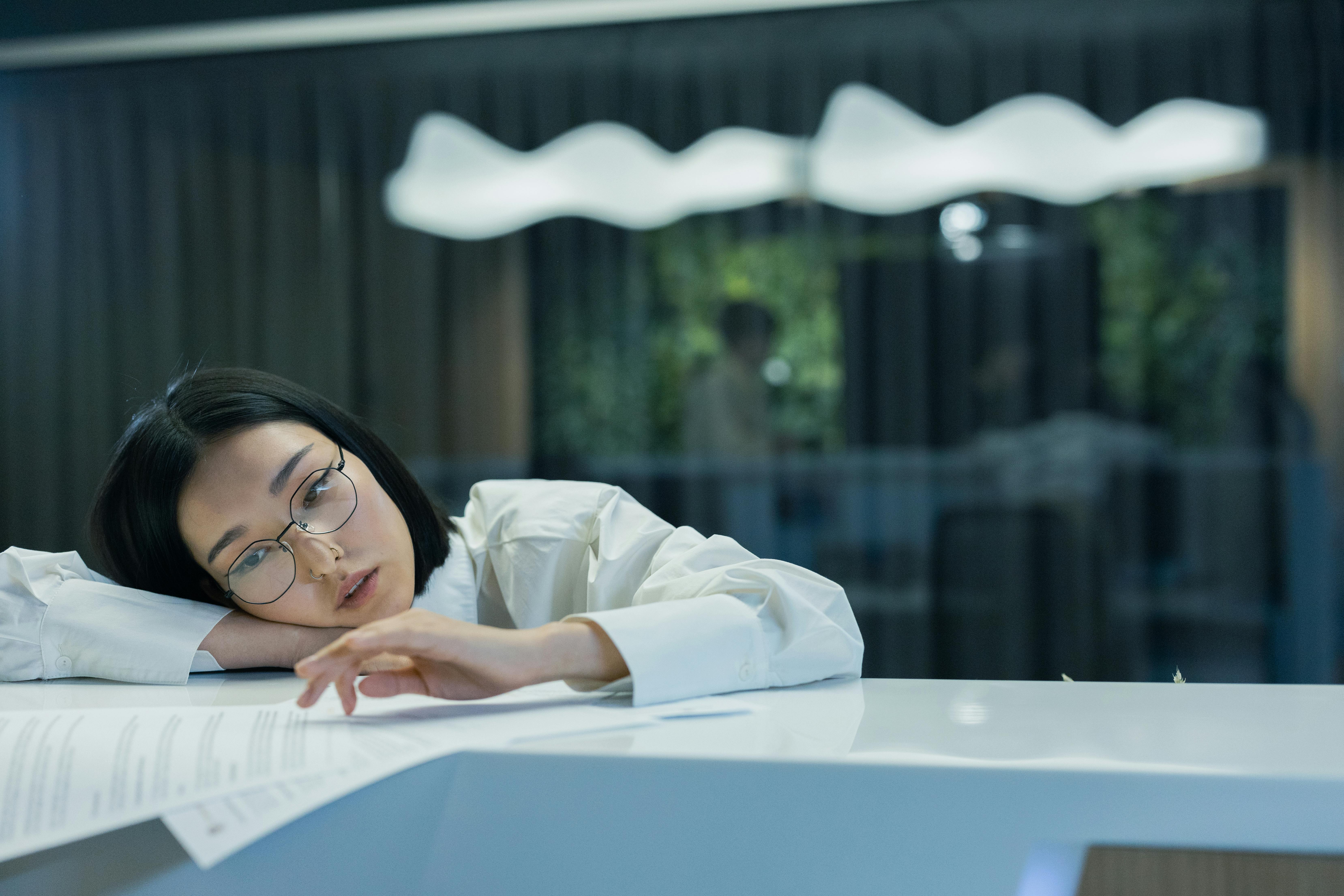 Asian woman in glasses rests her head on a counter with papers, showing signs of fatigue.
