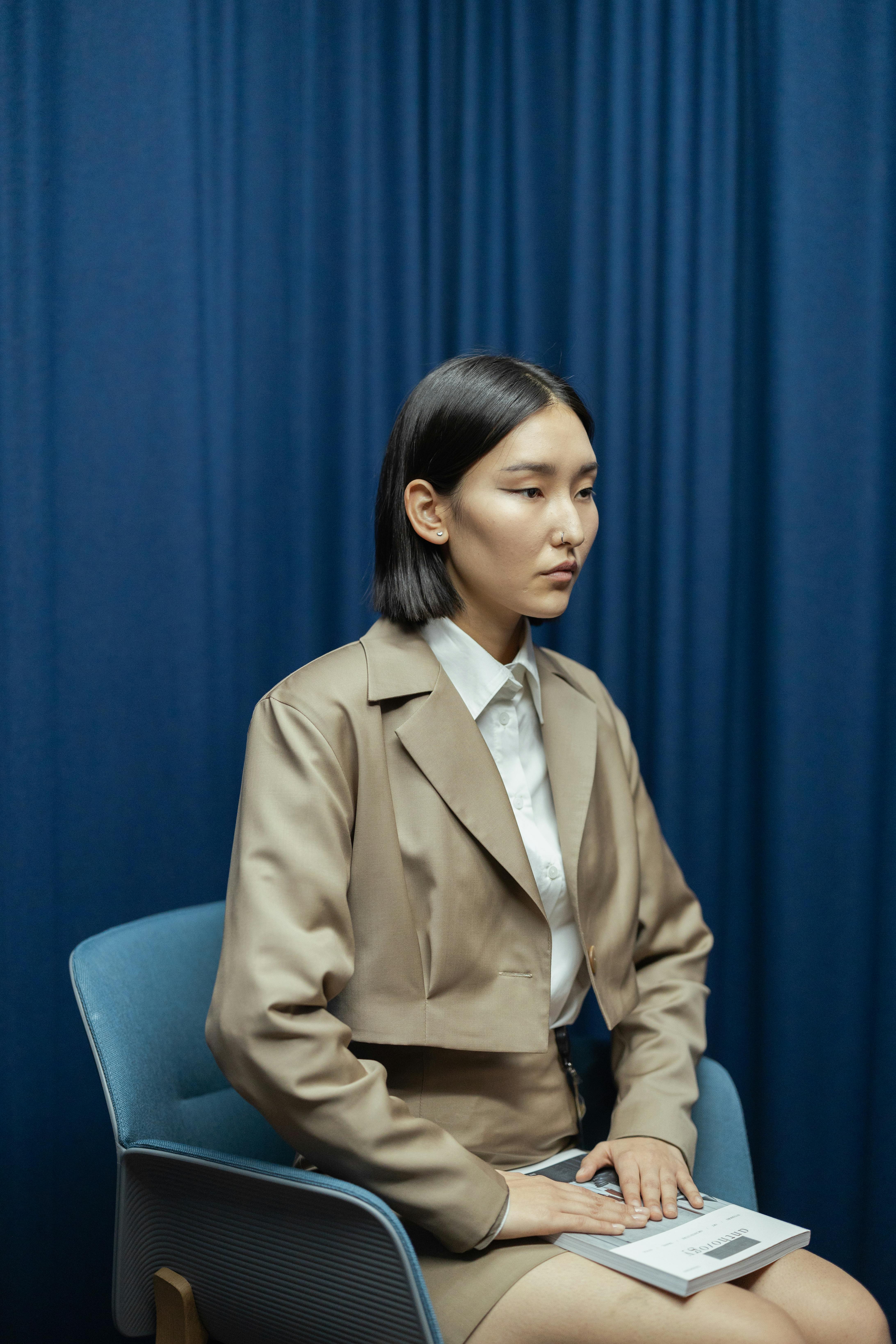 Portrait of a serious Asian woman sitting in business attire against a blue curtain backdrop.