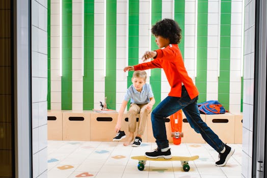 Two boys inside a colorful room, one skateboarding, the other sitting and observing.