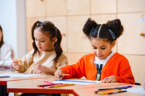 Free Two young girls creatively drawing with colored pencils in a bright classroom setting. Stock Photo