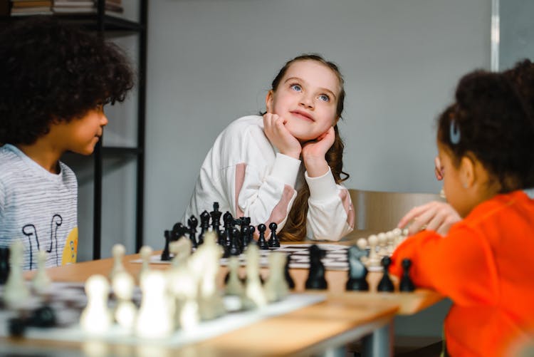 Girl In White Sweater Sitting Beside Kids Playing Chess Looking Up