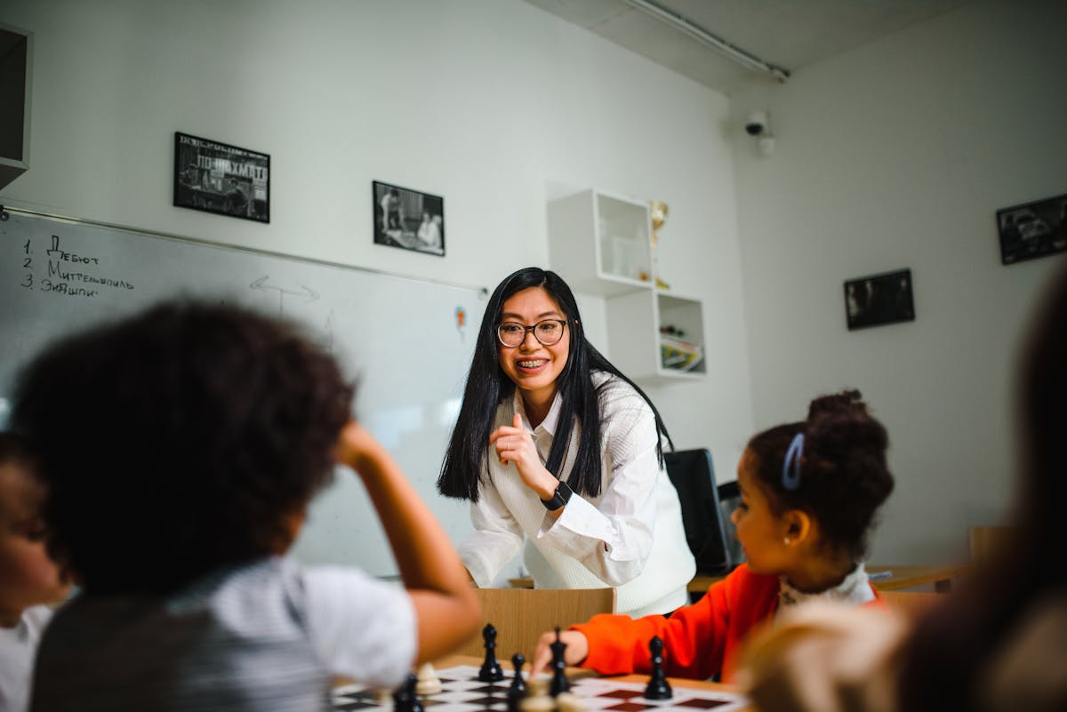 Teacher interacts with children during chess lesson, demonstrating critical thinking development through strategic gameplay