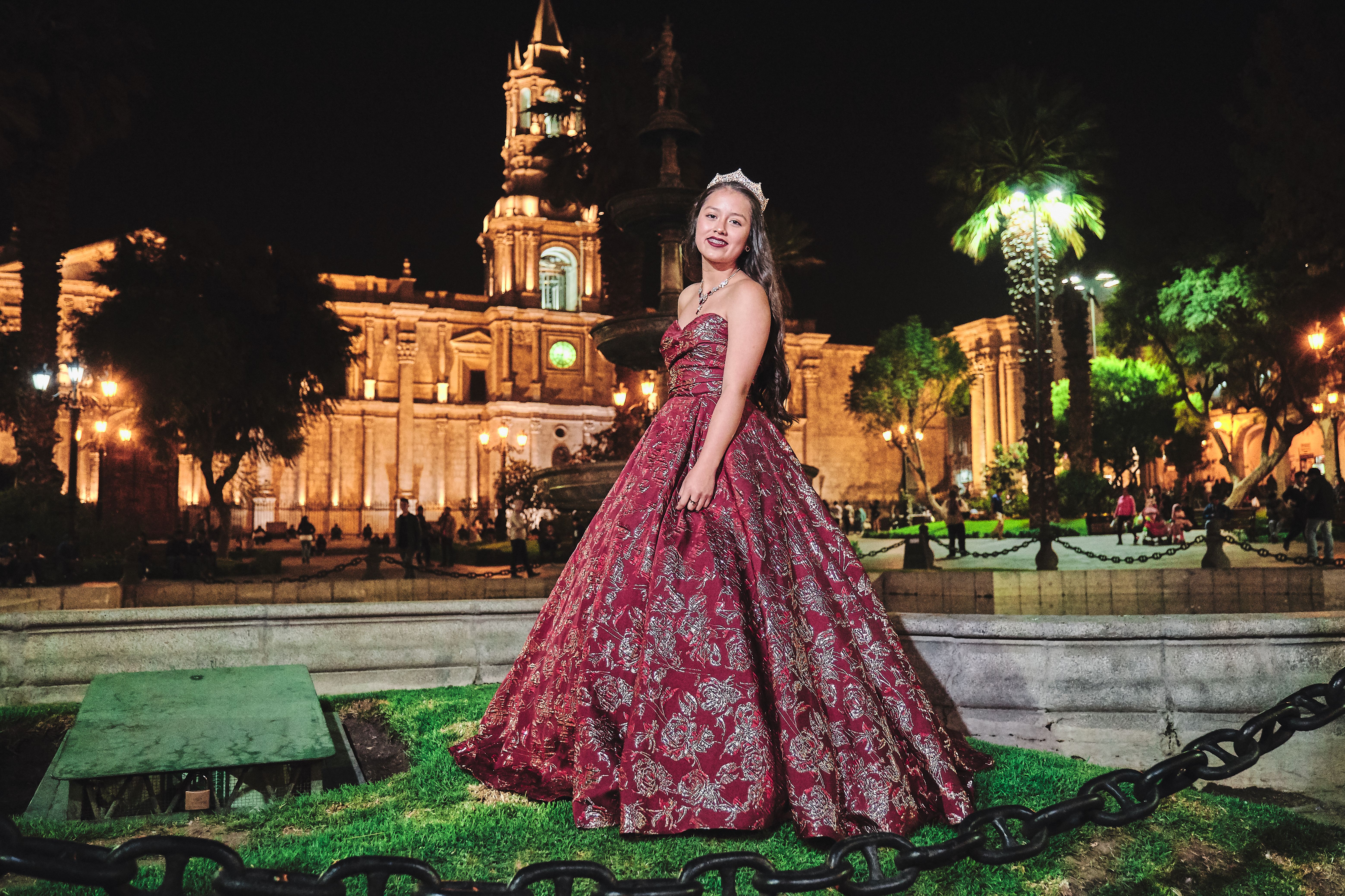 Young woman in red ball gown posing at night in a vibrant outdoor urban setting.