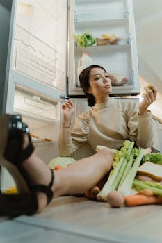 Asian woman sitting on the floor with vegetables near an open refrigerator, contemplating food choices.