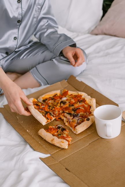 A person enjoys a fresh veggie pizza in bed with a warm cup of coffee.