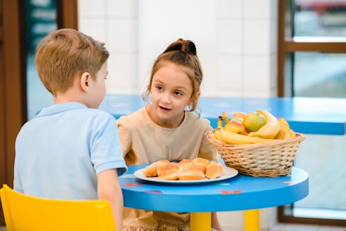 Girl and Boy Sitting Around Table With Food 