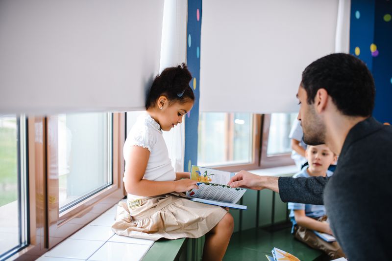 An adult supporting a child reading a book at a classroom table