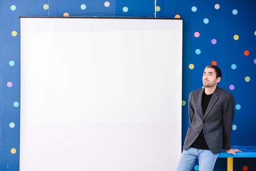 Adult man in a suit leaning against a table, with a whiteboard on a polka dot wall background.